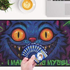 May include: A rectangular mousepad with a colourful illustration of a Cheshire Cat with orange eyes and a wide grin. The background features purple and green graffiti. A hand holds a fan of Pokemon cards.