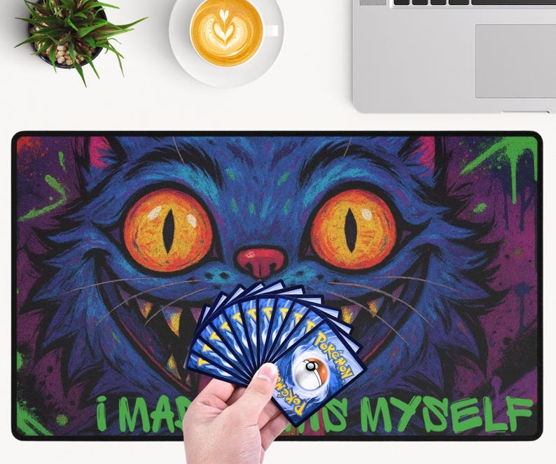 May include: A rectangular mousepad with a colourful illustration of a Cheshire Cat with orange eyes and a wide grin. The background features purple and green graffiti. A hand holds a fan of Pokemon cards.
