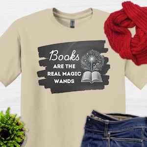Può includere: Una t-shirt beige con una grafica in bianco e nero che dice "Books are the real magic wands". La grafica presenta un libro con una bacchetta magica e stelle.