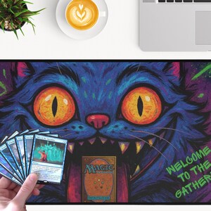 K&ouml;nnte beinhalten: Rechteckige Magic: The Gathering Spielmatte mit einem blauen Katzendesign, orangefarbenen Augen und scharfen Z&auml;hnen. Auf der Matte steht der Text "WELCOME TO THE GATHERING". Eine Hand h&auml;lt Magic-Karten.