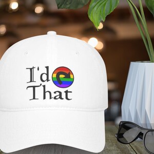 May include: White baseball cap with the text "I'd That" in black, featuring a rainbow-coloured arrow design. The cap is made of a durable fabric and has a curved brim. The design is centered on the front of the cap.