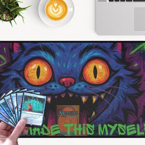 May include: A Magic the Gathering playmat with a blue cat design, orange eyes, and sharp teeth. The mat has the text "MADE THIS MYSELF" in green. A hand holds a stack of Magic the Gathering cards. The background is purple.