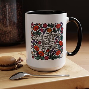 May include: White ceramic mug with a black handle and rim. The mug features a floral design with the text "Radicalized by Basic Decency" in a decorative font. The flowers are red, orange, purple, and pink.