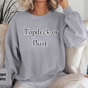 May include: A light grey sweatshirt with the text "Topdeck or Bust" in a black, decorative font. The sweatshirt has long sleeves and a crew neck. A smaller image of the sweatshirt is in the lower left corner, with the words "Sport Grey".
