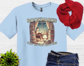 May include: Light blue t-shirt featuring a graphic of a person reading a book by a window. The text "Bookmarks They're for Quitters" is printed on the shirt.
