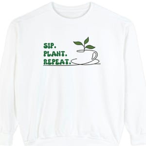 May include: White long-sleeve sweatshirt with a green graphic of a teacup with a plant growing out of it. The text "SIP. PLANT. REPEAT." is printed in green below the graphic.
