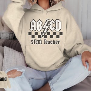 Peut inclure: Un sweat à capuche de couleur sable avec une poche avant et une capuche à cordon. Le devant présente un motif noir et blanc avec le texte "ABCD", un éclair et les mots "STEM Teacher". Le sweat à capuche est fait d'une matière douce.