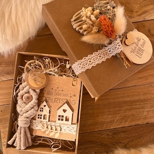Könnte beinhalten: Eine braune Geschenkbox mit einem herzförmigen Kranz aus getrockneten Blumen und einem Holzschild mit der Aufschrift "Hier steckst du Liebe drin". Die Box ist geöffnet und enthält eine kleinere Box mit einer Holzkarten mit der Aufschrift "Alles Liebe zum Geburtstag" und einem Makramee-Schlüsselanhänger mit einem Holzhaus-Anhänger.