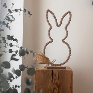 Könnte beinhalten: Dekorative Ostersilhouette eines Hasen aus beigefarbener Kordel, auf einem Holzblock stehend. Ein kleines Weizenbündel ist an der Basis befestigt. Der Hase ist neutral gefärbt, passend für die Frühlingsdekoration.