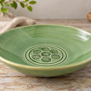 May include: A round, sage green ceramic bowl with a glossy finish. The center of the bowl features a recessed circular design containing seven small, embossed flower-like patterns arranged in a cluster, surrounded by concentric circular rings.