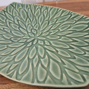 May include: A square ceramic plate in a muted sage green color featuring a raised, textured pattern of radiating petal-like shapes that originate from the center and extend outward toward the edges of the plate in a symmetrical design.
