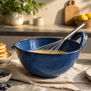 May include: A deep blue ceramic mixing bowl with a side handle and a pouring spout, containing a light yellow batter and a stainless steel wire whisk. The bowl has a textured, mottled finish and is positioned on a light-colored fabric cloth.