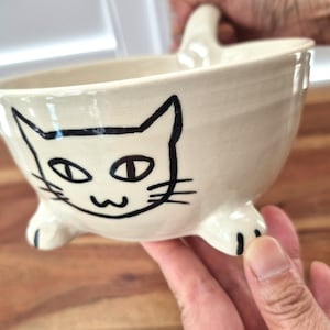 Puede incluir: Un cuenco de cerámica de color crema con un dibujo de líneas negras pintado a mano de una cara de gato en la parte frontal. El cuenco tiene tres pequeños pies con forma de pata de gato en la base y un asa en el borde, con un acabado brillante.