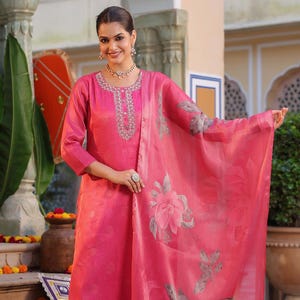 Puede incluir: Un vibrante conjunto de salwar kameez rosa. El atuendo incluye una túnica de manga larga con intrincados bordados alrededor del escote, pantalones a juego y una dupatta transparente con grandes estampados florales. La mujer lleva joyas y zapatos.