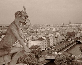Puede incluir: Una escultura de gárgola de piedra con una cara grotesca y cuernos, encaramada en una cornisa con vistas a una ciudad. La ciudad está en el fondo, con edificios y un río visibles.