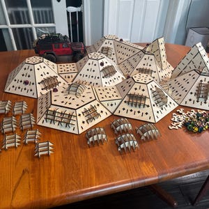 May include: A wooden game board featuring a variety of hexagonal pieces, some with small windows. The board is made from light wood and has a natural finish. There are also a number of small, colourful pieces scattered around the board.
