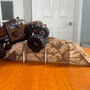 以下が含まれることがあります： 茶色とベージュの玩具トラックが黒い車輪で、茶色とベージュの迷彩模様のついた木製ランプを登っています。ランプは、互いに接続された複数の部品で構成されています。