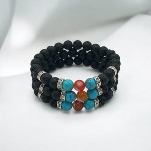 Könnte beinhalten: Ein dreireihiges Armband aus schwarzen Lavasteinen, Türkisen und Karneolen. Das Armband hat silberne Abstandshalter mit Strasssteinen.