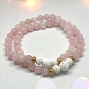 Könnte beinhalten: Zwei Armbänder mit Rosenquarzperlen, weißen Lavastein-Akzenten und goldfarbenen Abstandshaltern. Die Armbänder sind kreisförmig angeordnet und zeigen die zarte rosa Farbe des Quarzes.