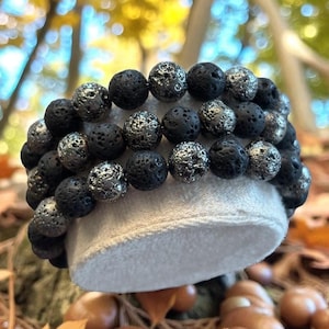 Könnte beinhalten: Drei Armbänder mit schwarzen und silbernen Lavasteinperlen. Die Perlen sind kugelförmig und strukturiert. Die Armbänder werden auf einer weißen Oberfläche präsentiert. Der Hintergrund ist eine verschwommene Naturkulisse mit Herbstfarben.