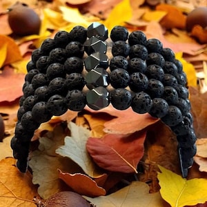 Könnte beinhalten: Ein schwarzes Armband mit Lavasteinperlen und facettierten Hämatitsteinen. Das Armband wird vor einem Hintergrund aus bunten Herbstblättern präsentiert, was seinen natürlichen und erdigen Stil unterstreicht. Das Design des Armbands ist schlicht, aber elegant.