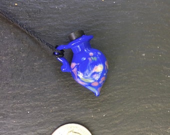 May include: A vibrant blue glass pendant necklace in the shape of a small pitcher. The pendant features colorful speckles and a black stopper. The necklace is strung on a black cord.