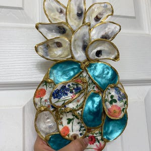 May include: A decorative pineapple made from oyster shells. The top is composed of natural oyster shells with gold trim. The body features teal shells and shells decorated with floral patterns. A unique coastal home decor item.