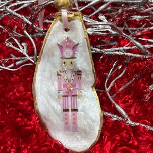 May include: A Christmas ornament featuring a pink and gold nutcracker soldier design on a white oyster shell. The shell is edged in gold and has a pink ribbon for hanging. The ornament is set against a red, textured background.