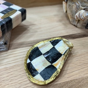 May include: An oyster shell dish featuring a hand-painted black and white checkered pattern on the interior surface. The irregular, scalloped edges of the shell are finished with a metallic gold trim, creating a distinct contrast against the monochrome pattern.