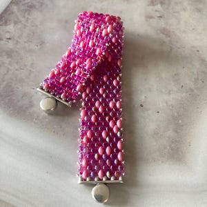 Può includere: Un braccialetto di perline caratterizzato da un motivo intrecciato di perline ovali rosa vivace e rosa tenue. Il braccialetto è rifinito con terminali in metallo argentato e una chiusura magnetica circolare, creando un aspetto strutturato.