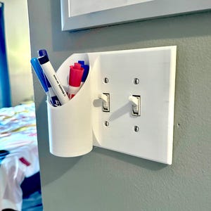 May include: White plastic wall mounted light switch with two toggle switches. A white plastic holder with a blue and pink marker and two blue pens is attached to the left side of the switch plate.