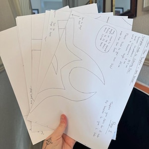 Könnte beinhalten: Ein Stapel weißer Papiervorlagen für einen Armschutz, mit handschriftlichen Notizen und Maßen in Zentimetern. Der Text enthält "Demo Show Template" und "Seite 1". Das Design zeigt geschwungene Linien und Skalierungsanweisungen.