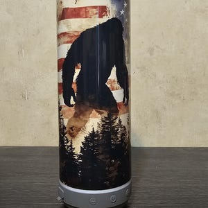 Puede incluir: Altavoz cilíndrico con un diseño que presenta una bandera estadounidense, una silueta de Bigfoot y una escena de bosque. El altavoz tiene una base gris con botones de control.