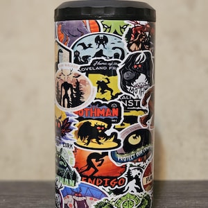 May include: A black and silver stainless steel tumbler covered in colorful stickers featuring various cryptids, including Mothman, the Dover Demon, and the Flatwoods Monster. The stickers feature text such as "Home of the Loveland Frog", "Protect Our Cryptids", and "Mothman 100".