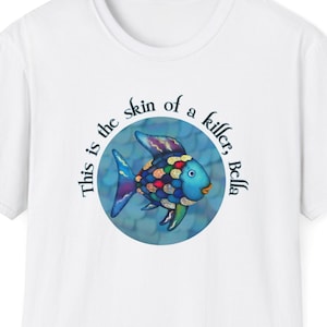 Puede incluir: Camiseta blanca con una ilustración de un pez colorido y el texto "This is the skin of a killer, Bella." El pez tiene escamas azules, moradas y amarillas. El texto está en un diseño circular alrededor del pez.