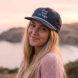 May include: A navy blue cap with the text "Your text or logo" in white. The cap features a white rope detail and a curved brim. The person in the image is wearing a pink sweater.