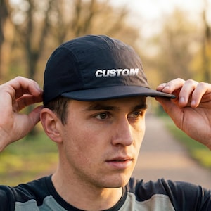 May include: Black cap with the word "CUSTOM" in white lettering. The cap has a curved brim and a mesh panel on the side. The person in the image is wearing a black and gray athletic shirt.