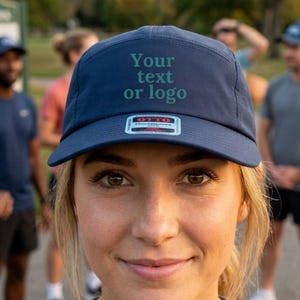 Puede incluir: Una gorra azul marino de cinco paneles con el texto Your text or logo impreso en verde oscuro en la parte frontal. La gorra incluye una pequeña pegatina rectangular en la visera con la marca Otto y texto adicional en blanco y rojo.