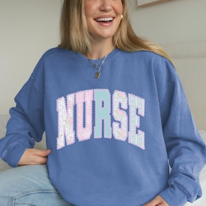 Peut inclure: Un sweat-shirt bleu à col rond avec le mot NURSE imprimé dans une grande police de style universitaire arquée. Les lettres ont un bord festonné et sont remplies d'un motif floral rose clair et bleu, créant un contraste net avec le tissu bleu uni du vêtement.