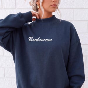 Peut inclure: Un sweat-shirt bleu marine à manches longues avec un col montant et des poignets côtelés. Le mot "Bookworm" est brodé en écriture cursive blanche au centre de la poitrine. Le vêtement présente une coupe décontractée avec des épaules tombantes.