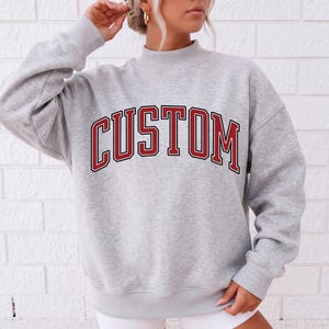 May include: A heather grey long-sleeve sweatshirt featuring a mock neck collar and ribbed cuffs. The word CUSTOM is printed across the chest in a bold, arched varsity-style font with red lettering and a thick black outline on the fabric.