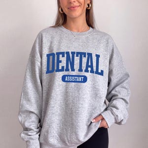 Op de afbeelding: Een grijs gemêleerd sweatshirt met ronde hals met het woord DENTAL in grote blauwe hoofdletters, en het woord ASSISTANT in het wit gedrukt in een blauwe horizontale pilvormige afbeelding direct onder de hoofdtekst.