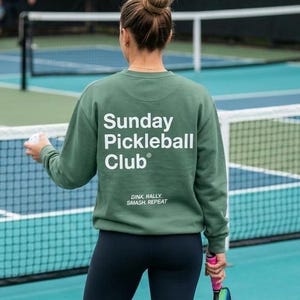Peut inclure: Une personne porte un sweat-shirt vert avec l'inscription "Sunday Pickleball Club" au dos, ainsi que la phrase "Dink, Rally, Smash, Repeat". Elle tient une raquette et une balle de pickleball sur un court, portant des leggings noirs et des baskets blanches.