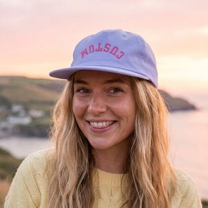 May include: A light purple baseball cap featuring the word CUSTOM embroidered in pink capital letters across the front panel. The cap has a structured crown, a curved brim, and a fabric strap closure at the back for an adjustable fit.