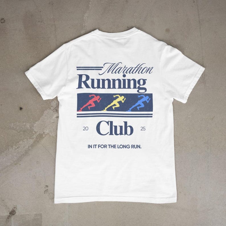 Puede incluir: Camiseta blanca con un dise&ntilde;o de Marathon Running Club. El dise&ntilde;o presenta el texto "Marathon Running Club" sobre un rect&aacute;ngulo azul con tres figuras de corredores en rojo, amarillo y azul. Tambi&eacute;n se incluye el texto "20 25" y "IN IT FOR THE LONG RUN".