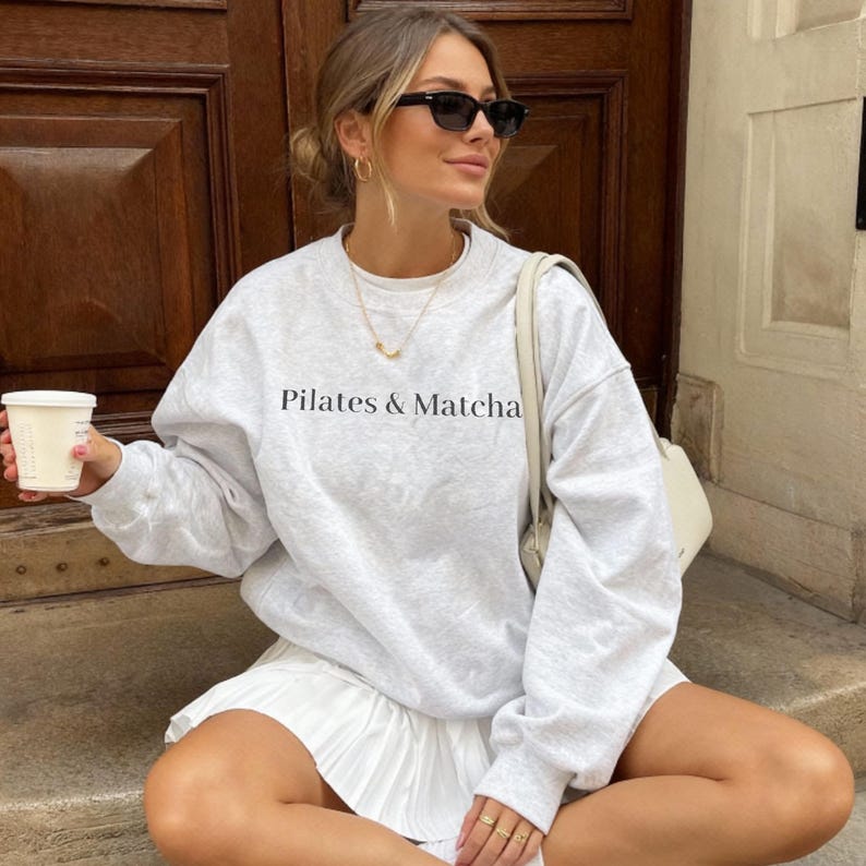Puede incluir: Una sudadera gris claro con el texto "Pilates & Matcha" en negro. La sudadera se combina con una falda plisada blanca. La persona lleva gafas de sol y sostiene una taza de caf&eacute;.