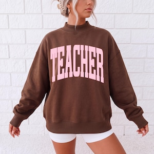Peut inclure: Un sweat-shirt à manches longues marron foncé avec un col montant. L'avant présente le mot TEACHER imprimé en grandes lettres majuscules rose clair arquées. Le vêtement a une coupe décontractée avec des poignets et un ourlet côtelés.