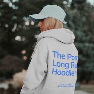 May include: A light gray hoodie with the text "The Post Long Run Hoodie" in blue on the back. The hoodie also has the text "Run Strong. Rest Well. Repeat." in blue. The person is wearing a light blue baseball cap.