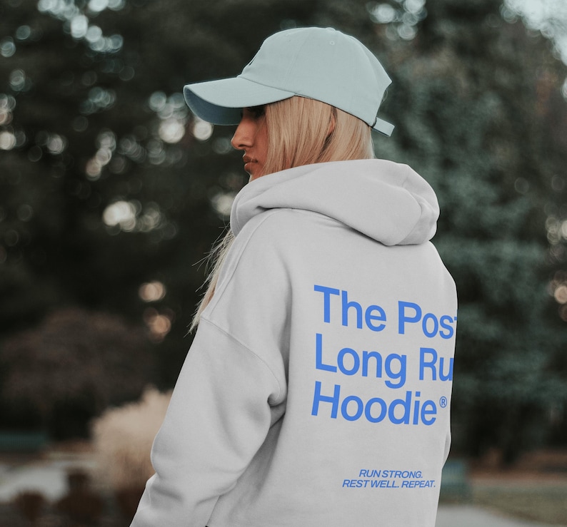 May include: A light gray hoodie with the text "The Post Long Run Hoodie" in blue on the back. The hoodie also has the text "Run Strong. Rest Well. Repeat." in blue. The person is wearing a light blue baseball cap.