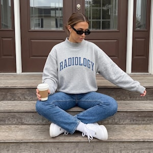 Könnte beinhalten: Ein graues Sweatshirt mit dem Wort "RADIOLOGY" in marineblauen Buchstaben. Das Sweatshirt wird mit blauen Jeans und weißen Turnschuhen kombiniert. Eine Person sitzt auf Holzstufen, hält eine Kaffeetasse und trägt eine schwarze Sonnenbrille.